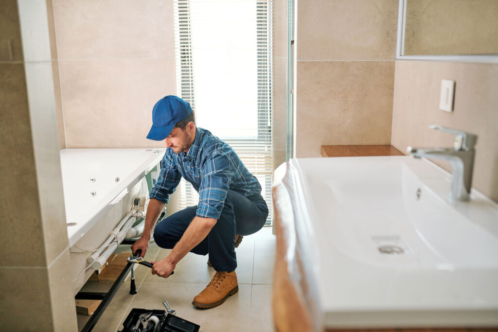 A bathroom in a stage of remodeling and a worker squatting down using a wrench.

